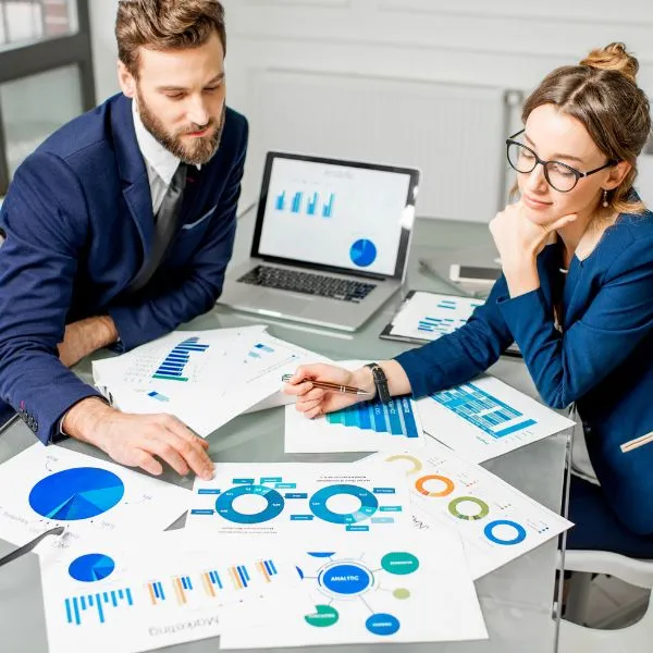 Two business professionals in an office setting reviewing a variety of colorful data charts, graphs, and infographics spread across a desk. A laptop in the background displays additional analytics.