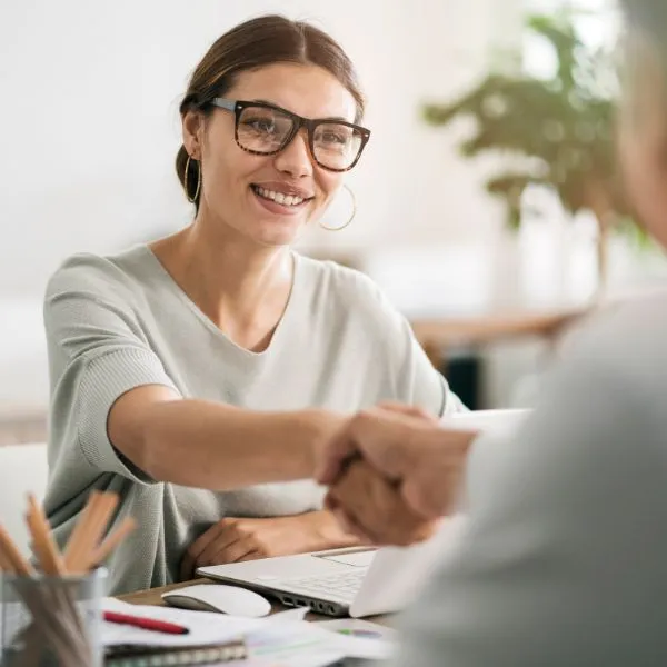 A smiling female business expert wearing glasses engages in a friendly handshake with a client over a desk.