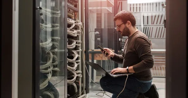 An IT technician uses a laptop and smartphone while troubleshooting a networked server rack in a data center.