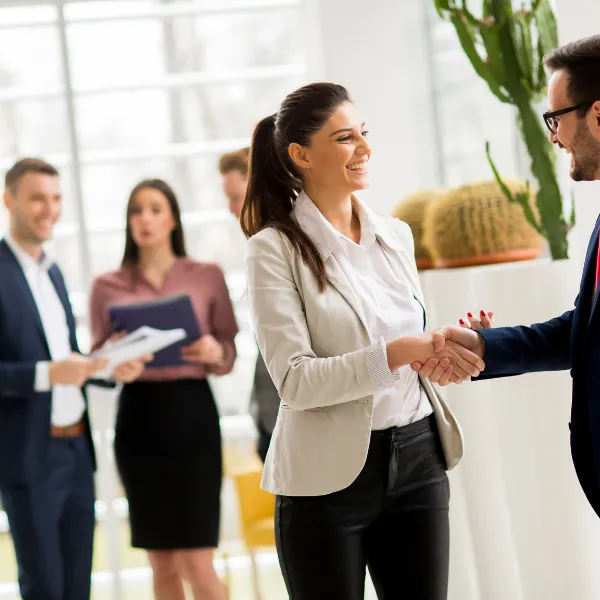 A diverse group of business professionals in a bright, modern office, featuring a woman in a blazer smiling while shaking hands with a colleague.