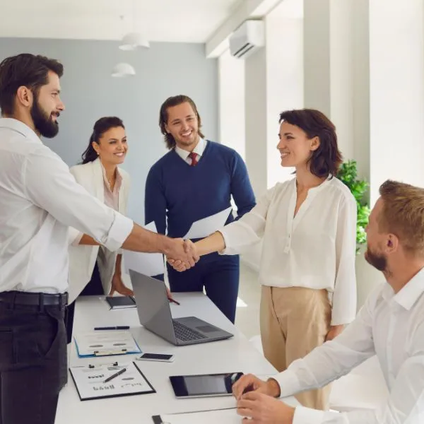 Two smiling professionals shaking hands across a conference table while three colleagues watch.